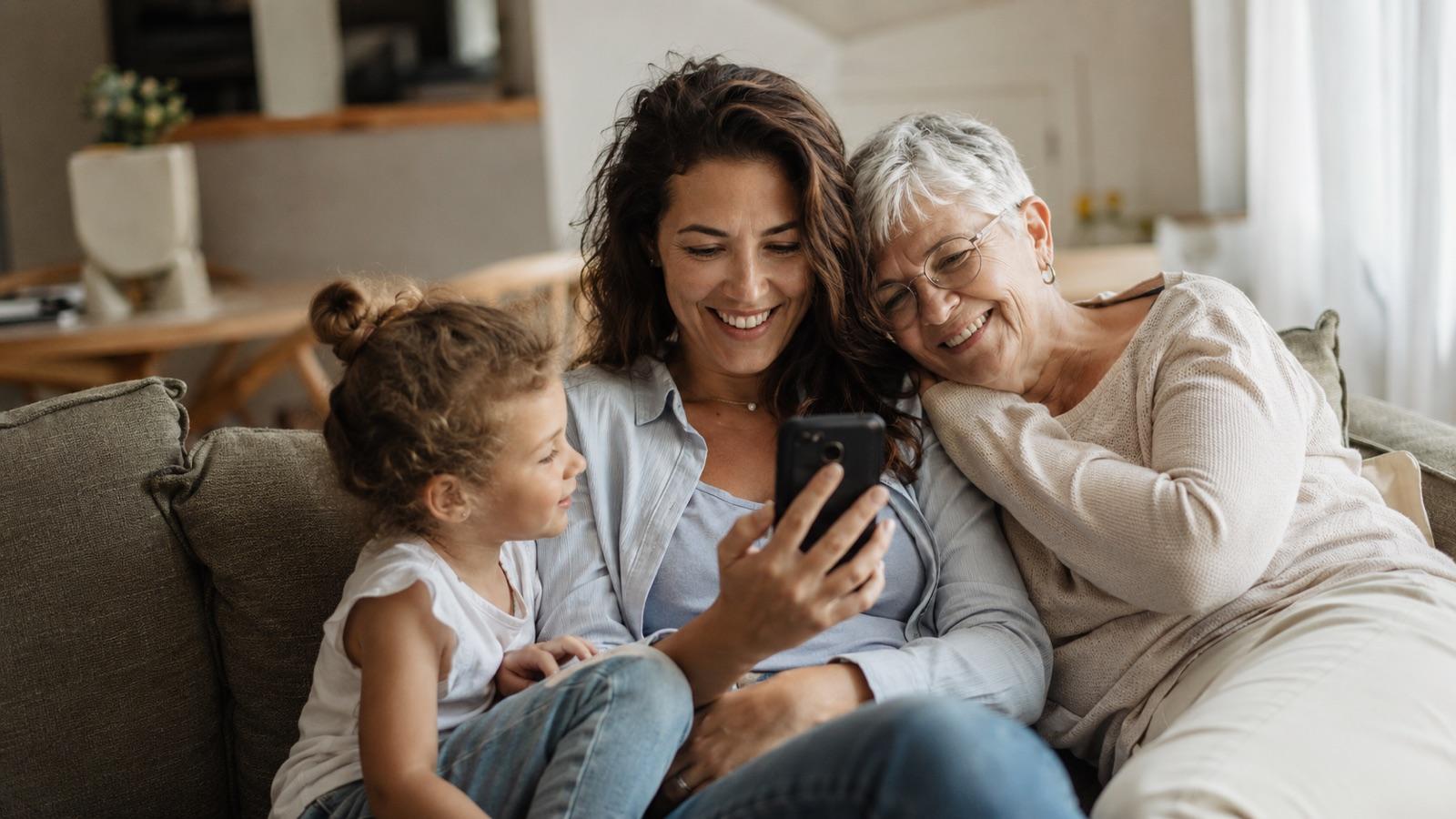 A mother, her young daughter, and a grandmother sitting together on a couch, smiling at a family memory on a phone